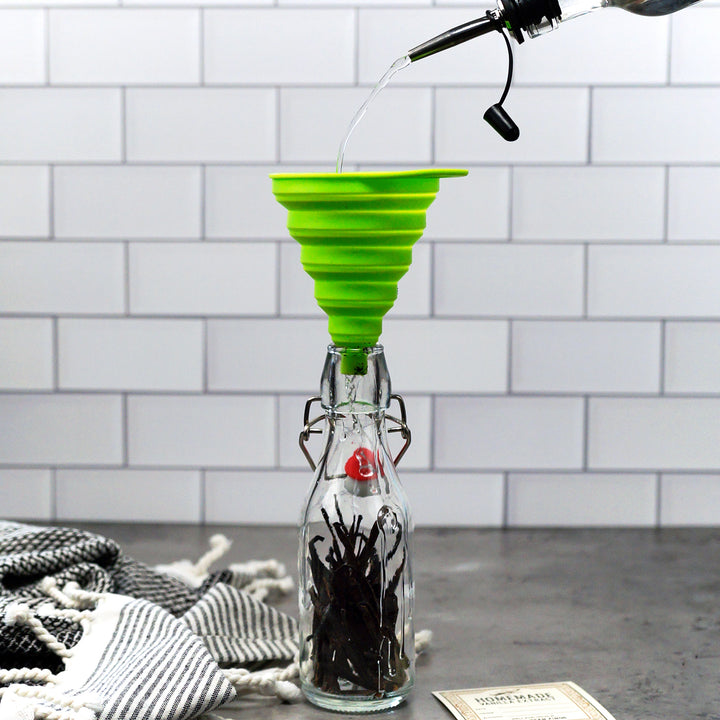 Green silicone funnel being used to pour liquid into a glass bottle with a white tiled wall background.