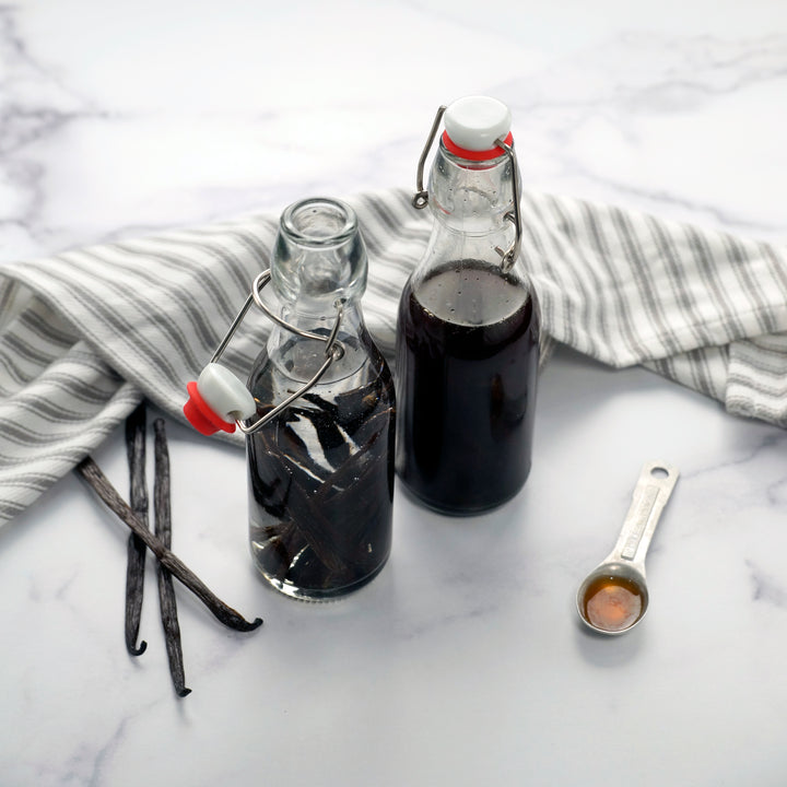 Two bottles of dark liquid with vanilla beans and a measuring spoon on a marble surface.