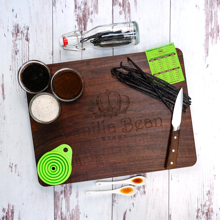 Wooden cutting board with vanilla beans, a knife, and small containers on a white wooden surface.